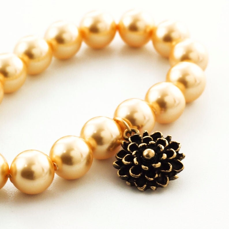 May include: A bracelet composed of smooth, round, metallic gold-toned beads arranged in a circular pattern. Attached to the strand is a single, dark bronze-colored metal charm shaped like a multi-petaled flower with a small central sphere.