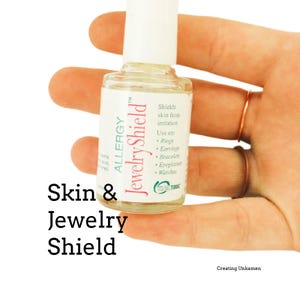 May include: A small clear bottle of Allergy Jewelry Shield with a white cap. The label features pink and green text stating it shields skin from irritation and is for use on rings, earrings, bracelets, eyeglasses, and watches. The bottle is held.