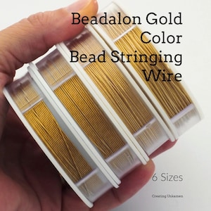 May include: Four spools of gold-coloured Beadalon bead stringing wire arranged in a row. The text on the image reads Beadalon Gold Color Bead Stringing Wire and 6 Sizes. The wire is wound onto clear plastic spools with white side discs.