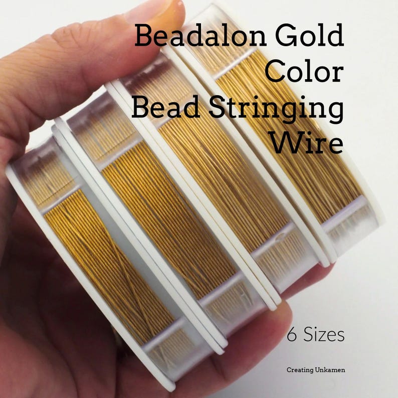 May include: Four spools of gold-colored Beadalon bead stringing wire arranged in a row. The text on the image reads Beadalon Gold Color Bead Stringing Wire and 6 Sizes. The wire is wound onto clear plastic spools with white side discs.