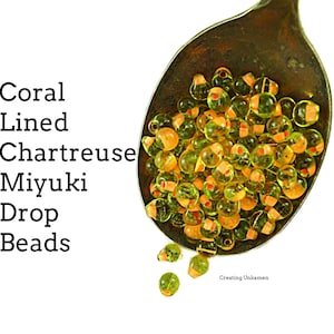 May include: A spoon filled with chartreuse green glass beads with a coral colored line around the center of each bead. The beads are called Miyuki Drop Beads.