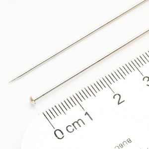May include: Two silver sewing pins, one with a rounded head, are placed on a white ruler marked in centimeters. The ruler shows measurements from 0 to 3 centimeters.