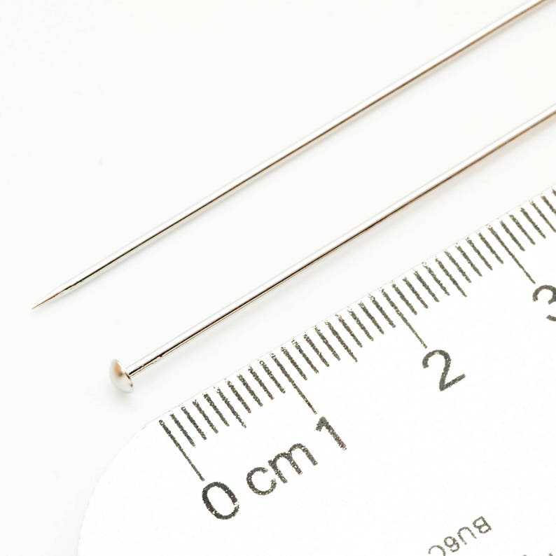 May include: Two silver sewing pins, one with a rounded head, are placed on a white ruler marked in centimeters. The ruler shows measurements from 0 to 3 centimeters.