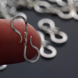 May include: Close-up of a silver-colored, twisted metal clasp. The clasp is in the shape of an elongated S, with a curved hook on each end. Other clasps are blurred in the background. The clasp is held between two fingers.