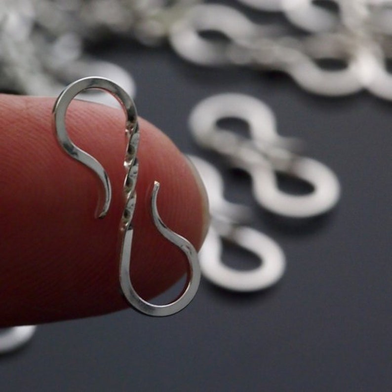 May include: Close-up of a silver-colored, twisted metal clasp. The clasp is in the shape of an elongated S, with a curved hook on each end. Other clasps are blurred in the background. The clasp is held between two fingers.