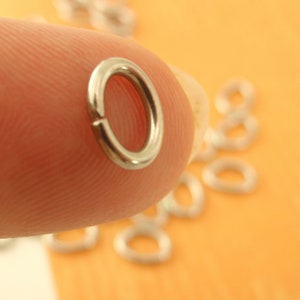 K&ouml;nnte beinhalten: Nahaufnahme eines silbernen Sprungrings, einer Art Schmuckzubeh&ouml;r, der von einem Finger gehalten wird. Der Ring ist rund und hat eine glatte Oberfl&auml;che. Mehrere andere Sprungringe sind im Hintergrund zu sehen, verstreut auf einer hellorangen Oberfl&auml;che.