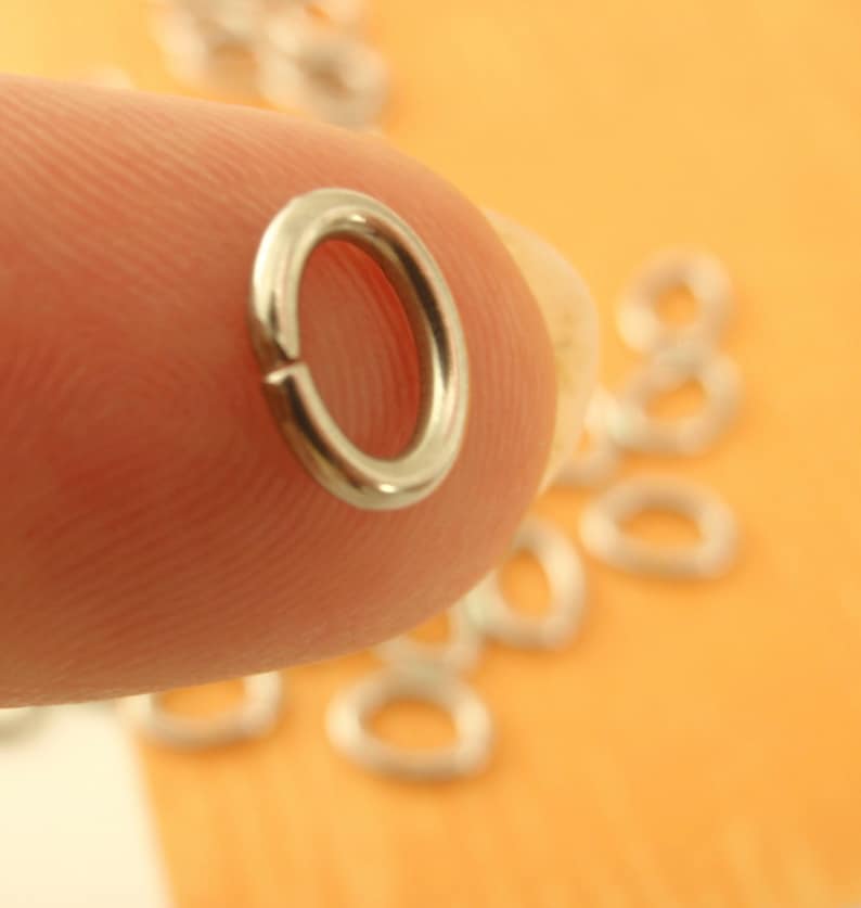 K&ouml;nnte beinhalten: Nahaufnahme eines silbernen Sprungrings, einer Art Schmuckzubeh&ouml;r, der von einem Finger gehalten wird. Der Ring ist rund und hat eine glatte Oberfl&auml;che. Mehrere andere Sprungringe sind im Hintergrund zu sehen, verstreut auf einer hellorangen Oberfl&auml;che.