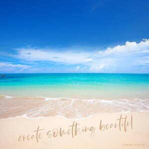 K&ouml;nnte beinhalten: Eine Strandszene mit blauem Himmel, wei&szlig;en Wolken und t&uuml;rkisfarbenem Wasser. Der Sand ist wei&szlig; und die Wellen schlagen an den Strand. Der Text "create something beautiful" ist in den Sand geschrieben.