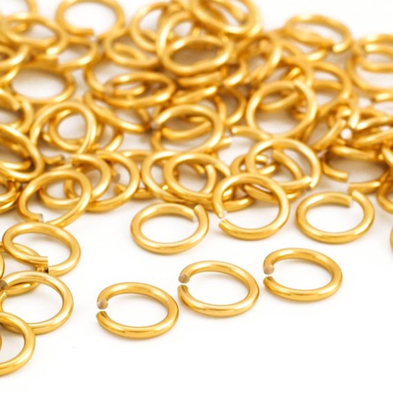 May include: A large pile of gold jump rings, a type of jewellery finding, are scattered on a white surface. The jump rings are small, circular, and have a smooth, shiny finish.