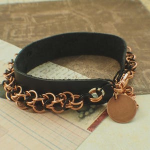 May include: A black leather bracelet featuring a chainmail border made of interlocking copper and black metal rings. The bracelet includes a circular copper-coloured metal charm attached to the side and secures with a metal clasp mechanism.