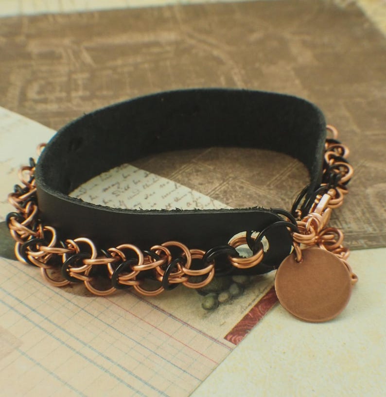 May include: A black leather bracelet featuring a chainmail border made of interlocking copper and black metal rings. The bracelet includes a circular copper-coloured metal charm attached to the side and secures with a metal clasp mechanism.