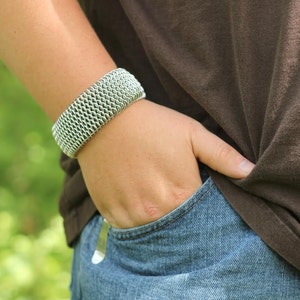 May include: A wide, silver-colored bracelet with a woven chainmail design. The bracelet is worn on a person's wrist, with the person wearing a brown shirt and blue jeans. The person's hand is in their jeans pocket.