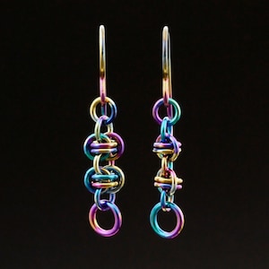May include: A pair of iridescent earrings with a chainmail design. The earrings feature a series of connected rings in shades of purple, blue, green, and gold. The earrings hang from a thin, elongated hook.