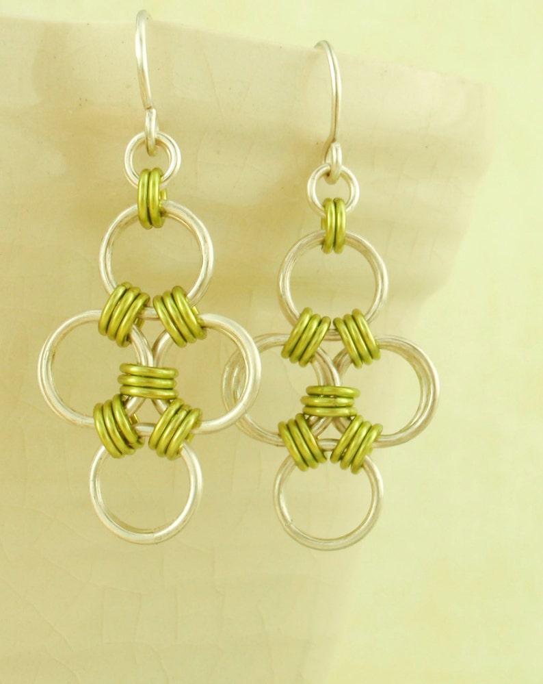 May include: Silver earrings with a green chainmail design. The earrings are made of multiple interlocking rings.