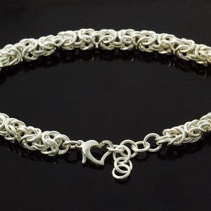 May include: A silver-colored bracelet with a woven chain design. The bracelet features a heart-shaped clasp and is displayed against a black background. This piece of jewelry is suitable for everyday wear.