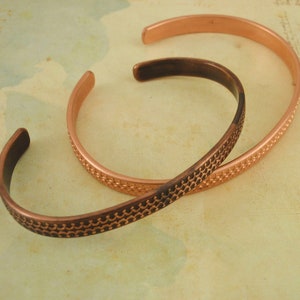 May include: Two copper bracelets with a textured pattern. The bracelets are stacked on top of each other.