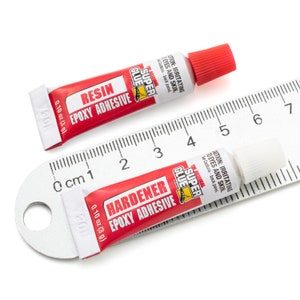 Puede incluir: Dos peque&ntilde;os tubos de adhesivo epoxi de resina, uno etiquetado como "Resina Adhesivo Epoxi" y el otro como "Dureza Adhesivo Epoxi". Ambos tubos son rojos y blancos con las palabras "Super Glue" impresas en ellos. Los tubos est&aacute;n sobre una regla con un fondo blanco.