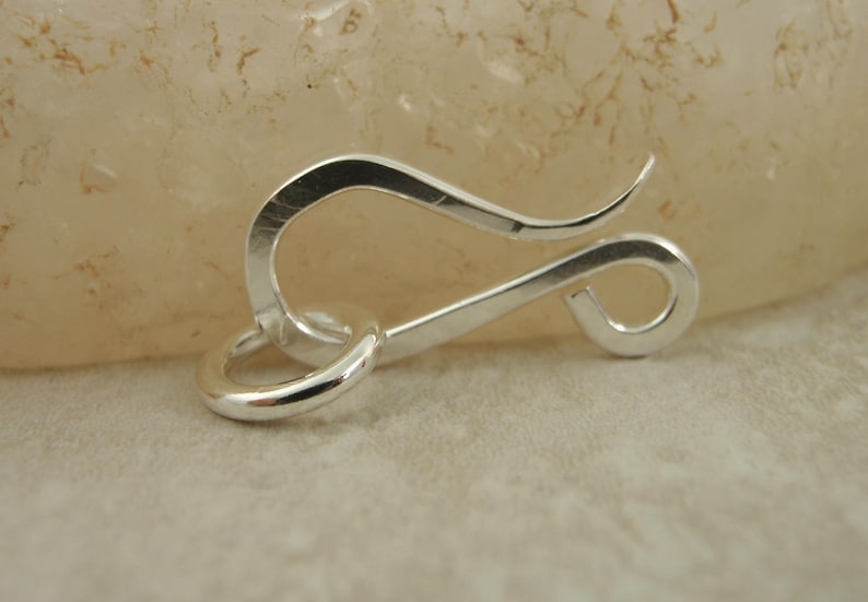 K&ouml;nnte beinhalten: Silberfarbener Metallverschluss mit einer Schlaufe an einem Ende und einem Haken am anderen. Der Verschluss ist zum Befestigen von Schmuck oder anderen Gegenst&auml;nden gedacht.