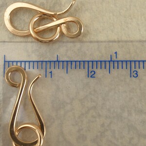 May include: Two gold-colored metal hook and eye jewelry clasps. One clasp is positioned horizontally above a ruler, while the other is placed vertically next to the ruler, showing a length of approximately 1 inch for the hook component.