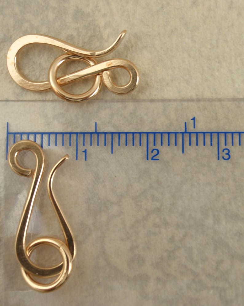 May include: Two gold-colored metal hook and eye jewelry clasps. One clasp is positioned horizontally above a ruler, while the other is placed vertically next to the ruler, showing a length of approximately 1 inch for the hook component.