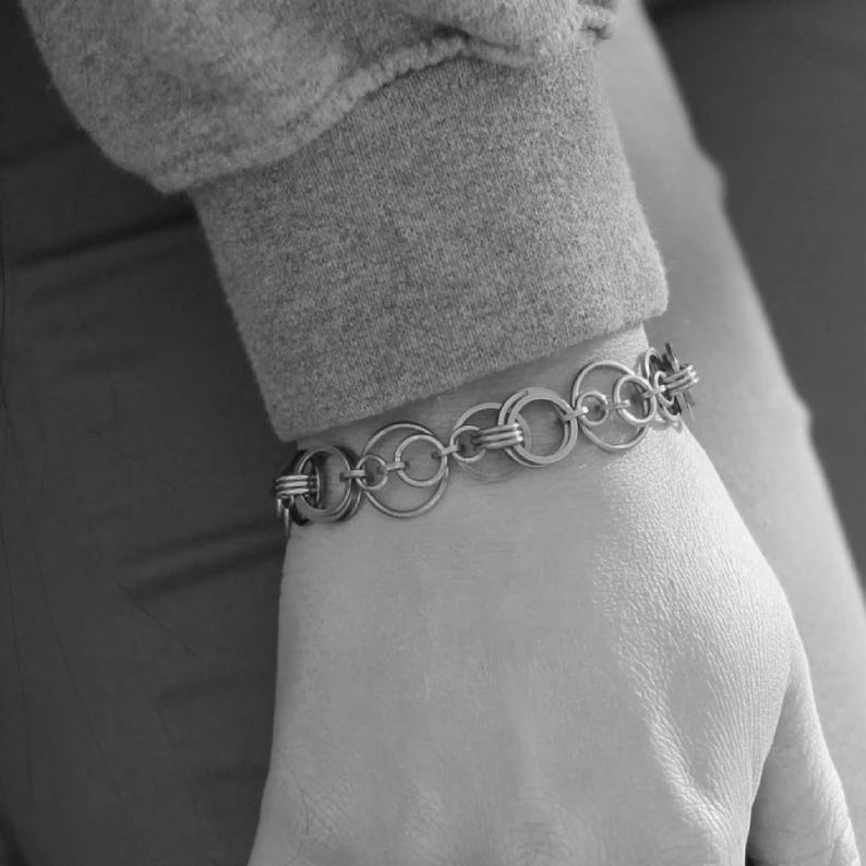 May include: A silver bracelet composed of linked circles and smaller rings. The bracelet is worn on a wrist, with a grey sleeve in the background. The design is simple and elegant, suitable for everyday wear.