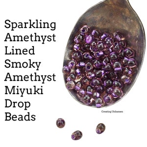 Puede incluir: Primer plano de una cuchara de plata llena de pequeñas cuentas de vidrio púrpura iridiscente. Las cuentas se describen como "Cuentas Miyuki de gota de amatista ahumada, amatista brillante, con borde de amatista". Tres cuentas están dispersas sobre un fondo blanco.