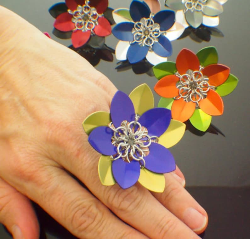 May include: A purple and yellow flower-shaped ring with a silver center. The ring is on a person's hand.