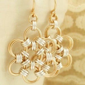 May include: A pair of dangle earrings featuring a circular flower-like pattern constructed from interlocking gold and silver-toned metal rings. Each earring hangs from a simple gold-toned hook, showcasing a geometric chainmaille design.
