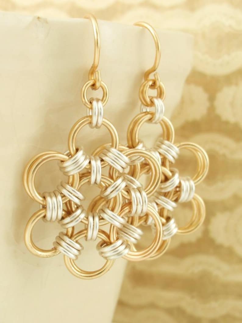May include: A pair of dangle earrings featuring a circular flower-like pattern constructed from interlocking gold and silver-toned metal rings. Each earring hangs from a simple gold-toned hook, showcasing a geometric chainmaille design.