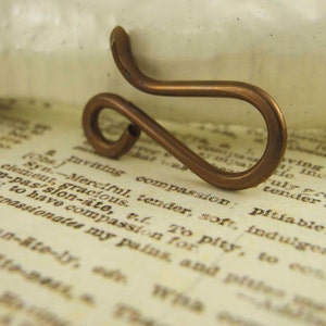 Può includere: Un elemento di bigiotteria artigianale color rame. Il pezzo di metallo è piegato in una forma unica e astratta con un'estremità ad anello e un design curvo e aperto. L'elemento è posto su uno sfondo di pagine di libri d'epoca.