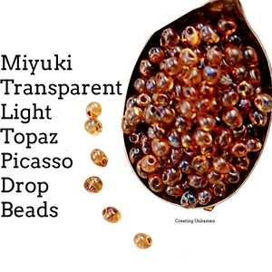 May include: A spoon filled with small, amber-colored glass beads. The beads are teardrop shaped and have a slight iridescent sheen. The text "Miyuki Transparent Light Topaz Picasso Drop Beads" is visible in the image.