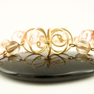 May include: A gold and copper wire necklace with clear quartz beads. The necklace features a decorative, intricate design with a repeating pattern of swirls and loops.