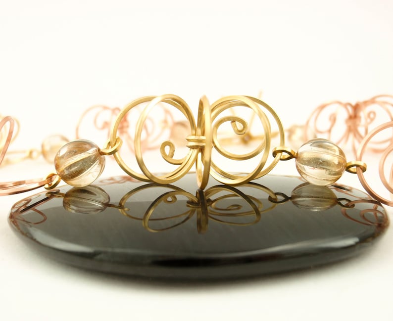 May include: A gold and copper wire necklace with clear quartz beads. The necklace features a decorative, intricate design with a repeating pattern of swirls and loops.