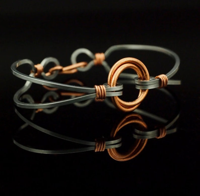 May include: A bracelet constructed from dark grey metal wire, featuring a central circular copper ring element. The dark grey wire is shaped into loops and secured with thin copper wire wrappings at multiple points along the band structure.