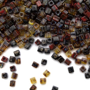 May include: A pile of small, square glass beads in shades of brown, black, and red. The beads are scattered on a white surface.