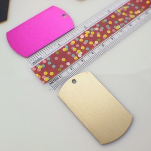 May include: Two blank metal dog tags, one pink and one gold, with a hole at the top for a ring. The tags are lying on a white surface next to a ruler with a red and white polka dot pattern.