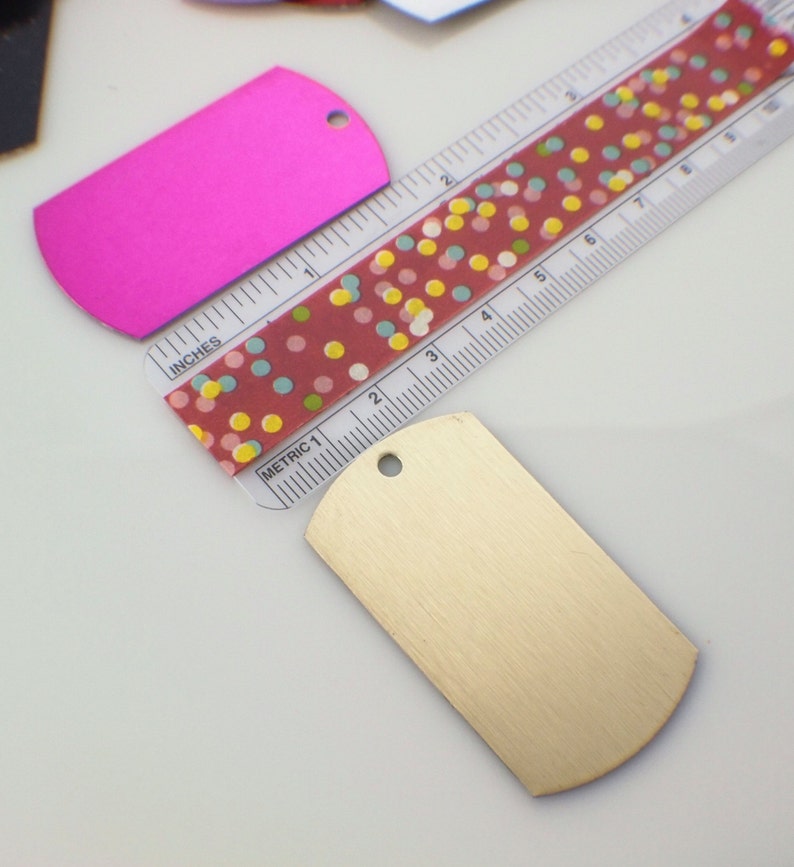 May include: Two blank metal dog tags, one pink and one gold, with a hole at the top for a ring. The tags are lying on a white surface next to a ruler with a red and white polka dot pattern.