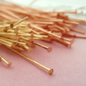 Op de afbeelding: Close-up van een verzameling goud- en koperkleurige kopspelden. De spelden hebben een rechte, slanke schacht met een platte, ronde kop. De spelden zijn verspreid over een roze oppervlak.