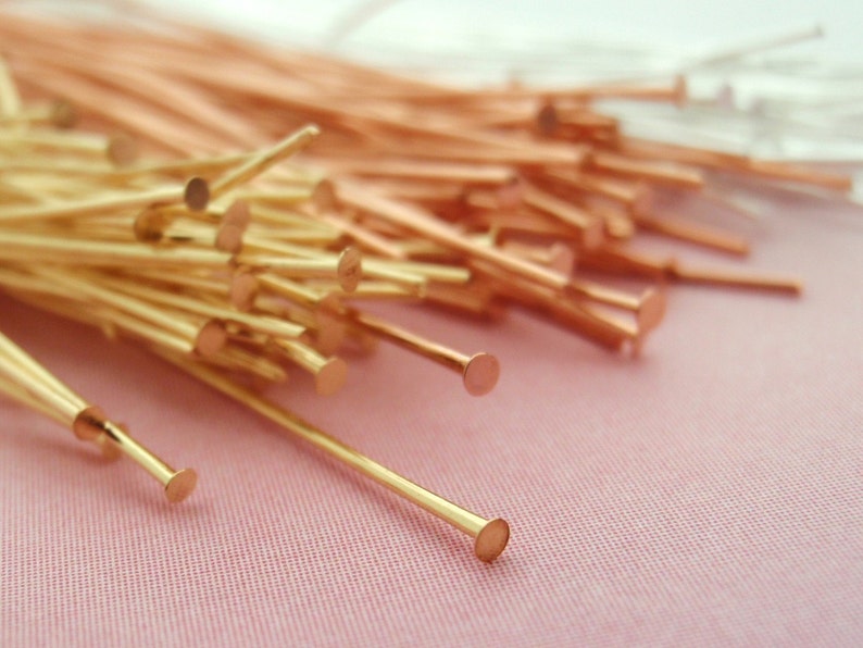 Op de afbeelding: Close-up van een verzameling goud- en koperkleurige kopspelden. De spelden hebben een rechte, slanke schacht met een platte, ronde kop. De spelden zijn verspreid over een roze oppervlak.