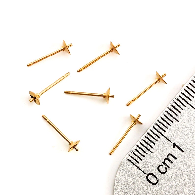 May include: Six gold-toned metal earring posts with flat circular pads for attaching components. Each post features a thin stem and a small peg at the center of the pad. A portion of a metal ruler is visible, showing a scale in centimeters.