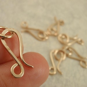 May include: A gold-toned metal hook clasp with a curved, elongated shape and a circular loop at one end. The clasp features a smooth, polished finish and is designed for jewelry making, with several identical pieces visible in the background.