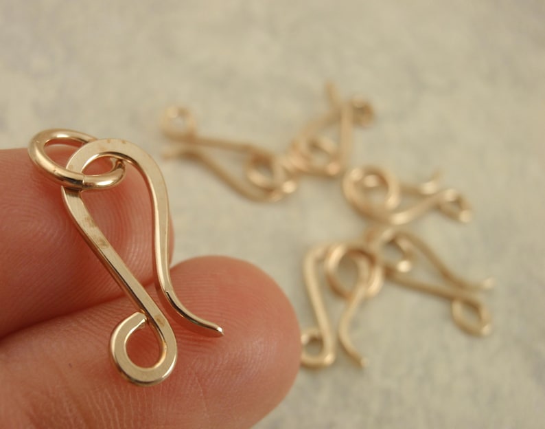 May include: A gold-toned metal hook clasp with a curved, elongated shape and a circular loop at one end. The clasp features a smooth, polished finish and is designed for jewelry making, with several identical pieces visible in the background.