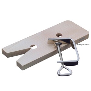 May include: A light-colored wooden bench pin with a V-shaped cutout and two circular holes, accompanied by a silver-colored metal C-clamp with a threaded screw mechanism. The text "Creating Unkamen" is printed on the surface of the wood.