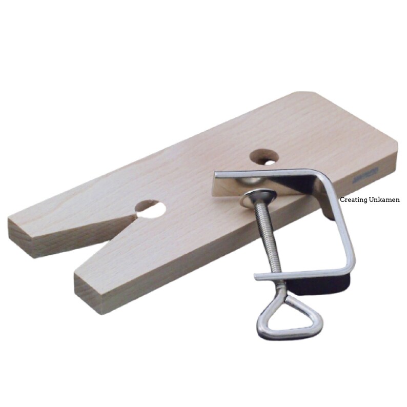 May include: A light-colored wooden bench pin with a V-shaped cutout and two circular holes, accompanied by a silver-colored metal C-clamp with a threaded screw mechanism. The text "Creating Unkamen" is printed on the surface of the wood.