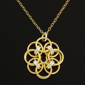 Op de afbeelding: Een goudkleurige ketting met een bloemvormige hanger. De hanger bestaat uit in elkaar grijpende gouden ringen met zilverkleurige accenten. De ketting is ook goudkleurig, tegen een zwarte achtergrond.
