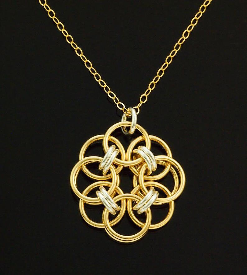 Op de afbeelding: Een goudkleurige ketting met een bloemvormige hanger. De hanger bestaat uit in elkaar grijpende gouden ringen met zilverkleurige accenten. De ketting is ook goudkleurig, tegen een zwarte achtergrond.