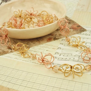 May include: A gold and rose gold wire necklace with delicate flower-shaped charms and clear glass beads. The necklace is displayed on a sheet of music.