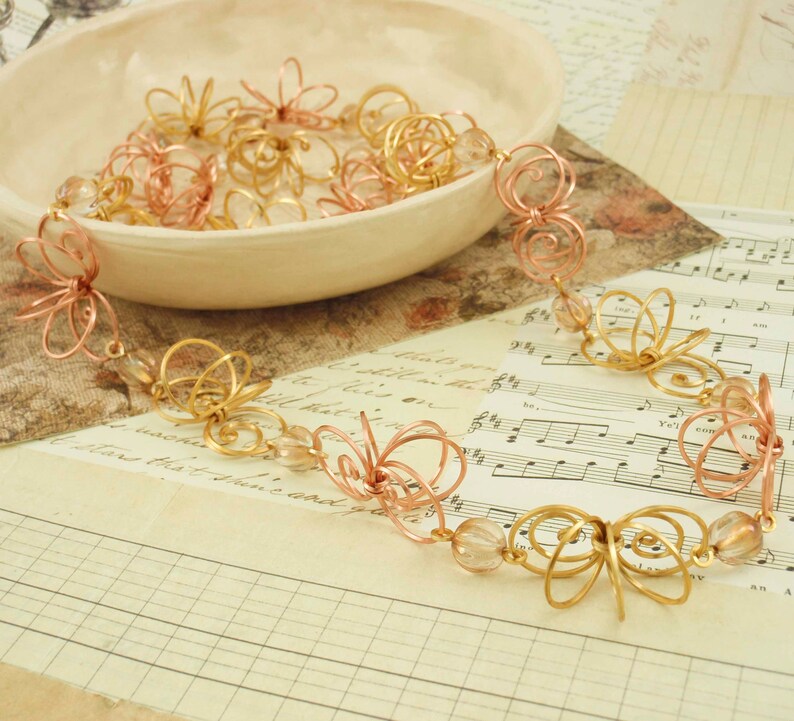 May include: A gold and rose gold wire necklace with delicate flower-shaped charms and clear glass beads. The necklace is displayed on a sheet of music.