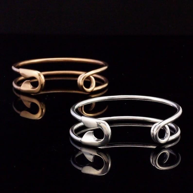 K&ouml;nnte beinhalten: Zwei Armb&auml;nder aus Metall, eines golden und eines silbern, in Form von Sicherheitsnadeln. Die Armb&auml;nder befinden sich auf einem schwarzen Hintergrund.