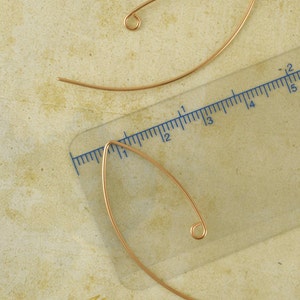 May include: Two curved, gold-toned wire earring components with a loop at one end. A clear plastic ruler is positioned alongside the lower earring, showing a length of approximately 1.5 inches from the pointed tip to the curved base.